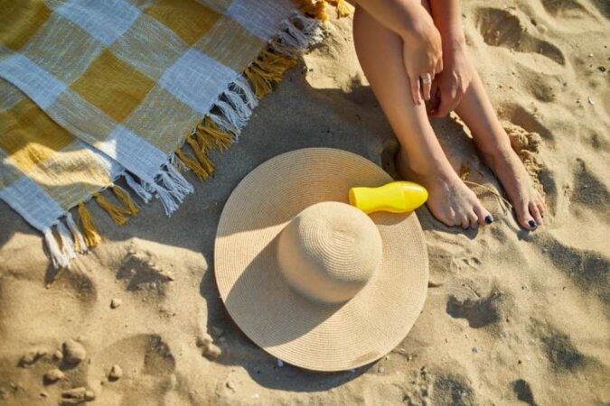 Une femme assise sur le sable chaud présente ses jambes et ses pieds nus près d'un chapeau et d'un tube de crème solaire, éléments clés d'une routine beauté estivale.